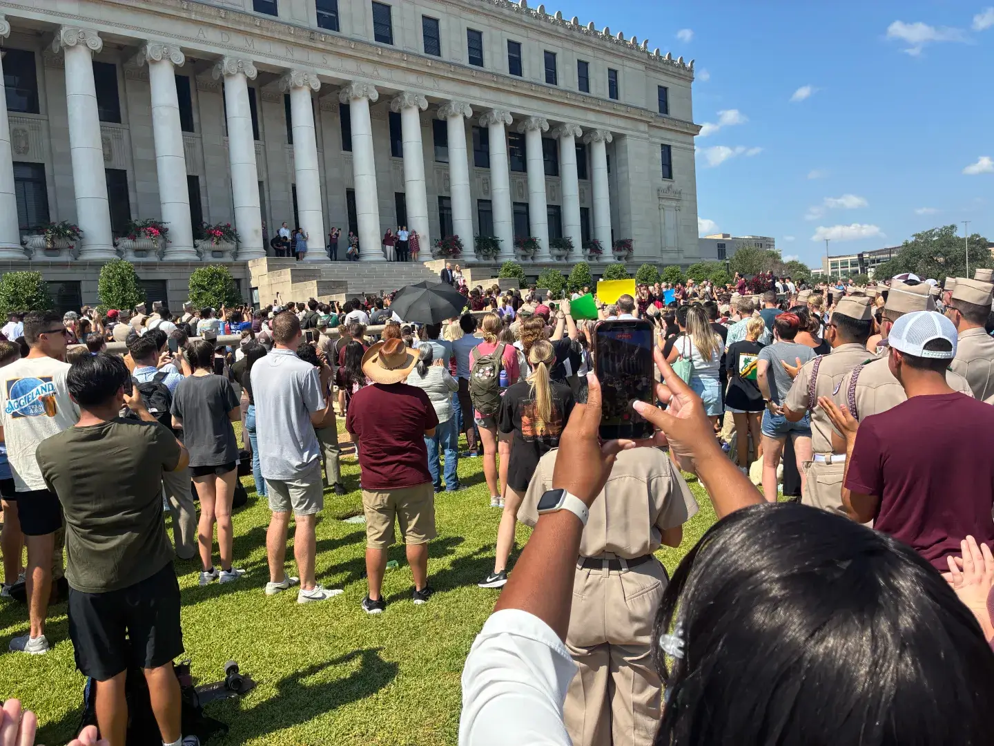 A large crowd gathers outdoors near a historic building on a sunny day.